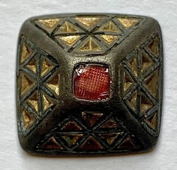 photo of a pyramid shaped gold-coloured object with decorative patterns of triangles on the sides and a square piece of red glass or gemstone at the peak. 